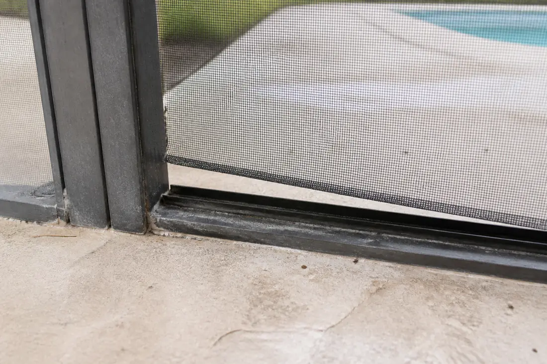 A torn screen panel at the base of a pool enclosure beside a door